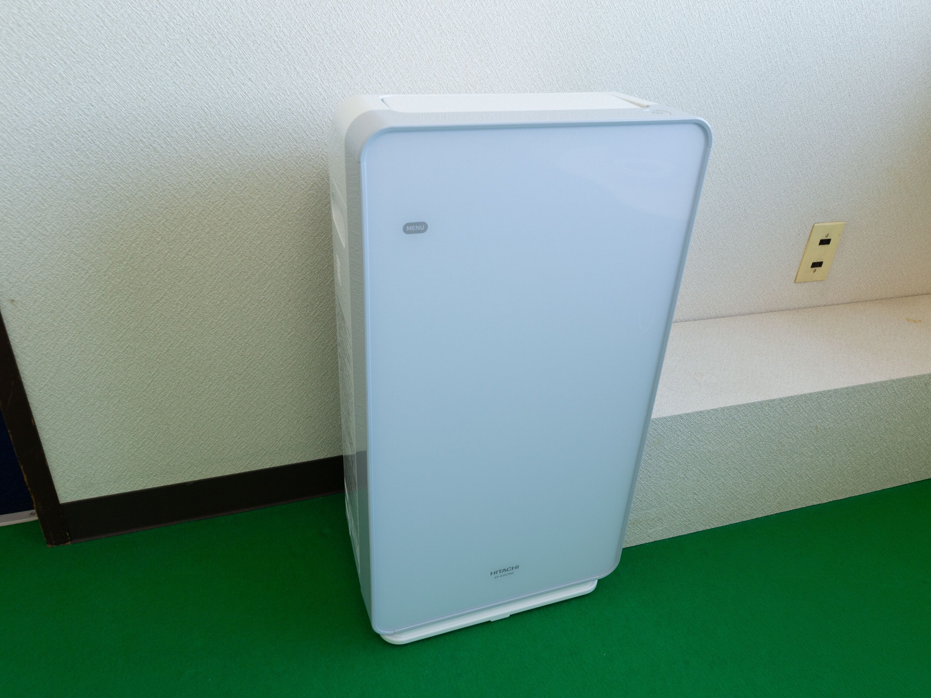 A white, rectangular device with a small control panel on the top, standing on a green floor against a white wall.