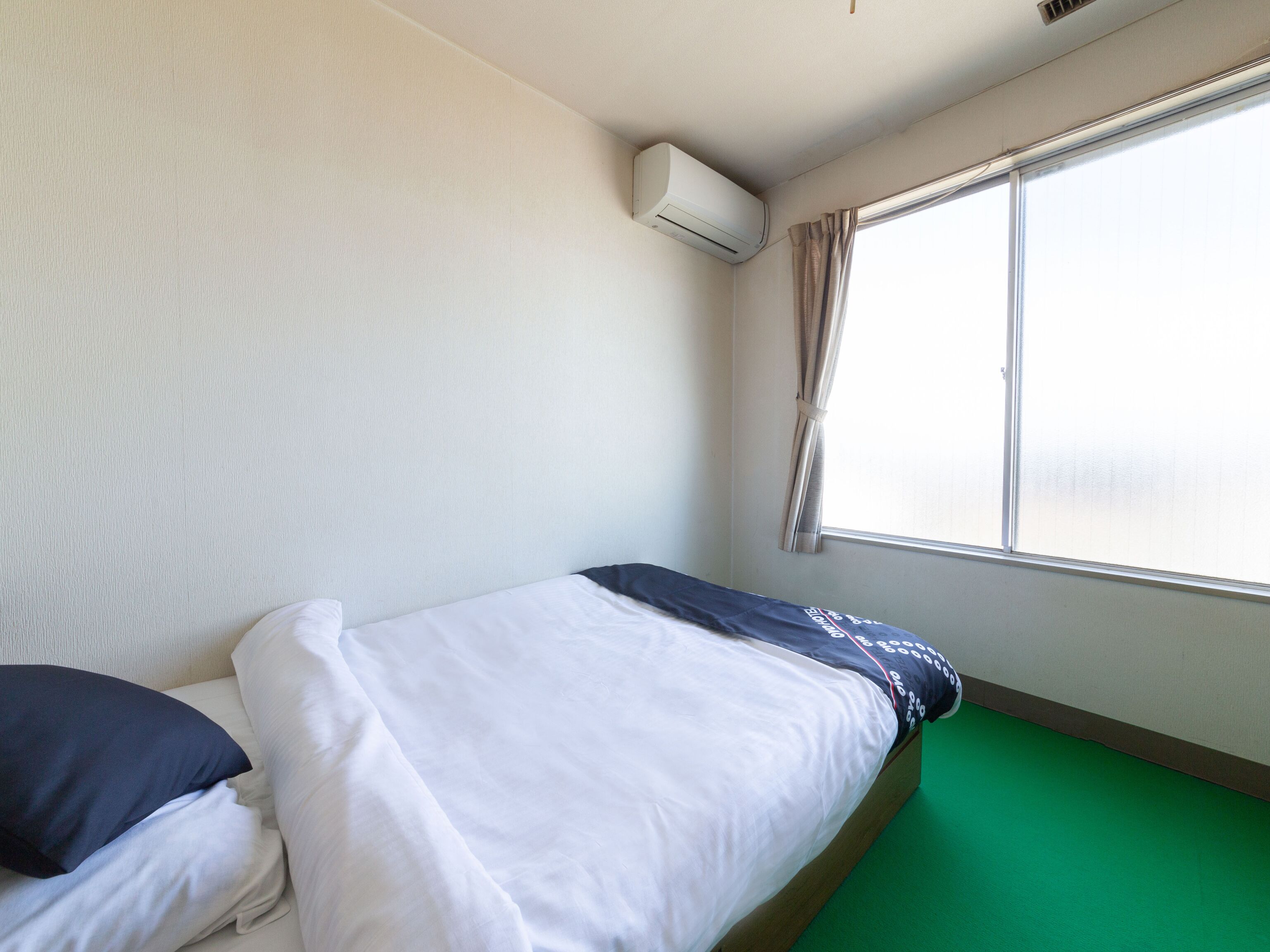 A single bed with white bedding and a green mattress, a window with curtains, and an air conditioning unit on the wall.