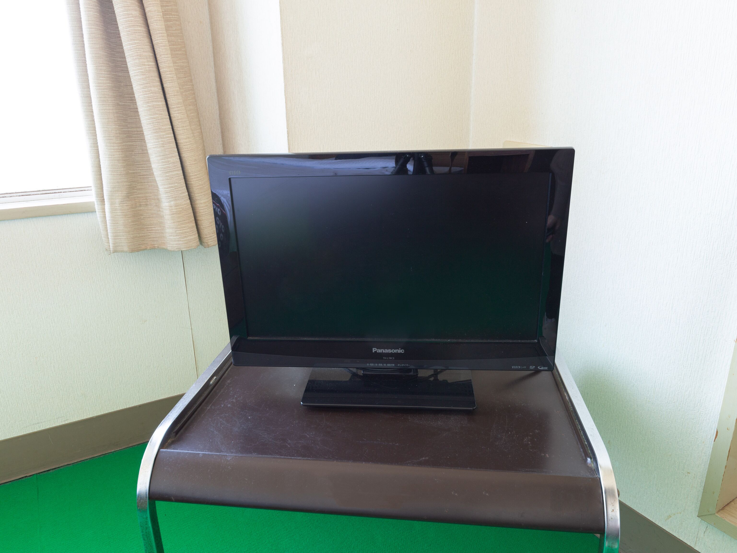 A Panasonic TV on a black stand in a room with a green floor and beige curtains.