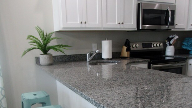 A kitchen with white cabinets, a granite countertop, a stainless steel microwave, and three light blue stools. - Stay & Play: Pet Friendly Extended Stay for Sports Travel
Covered Parking (Myrtle Beach)