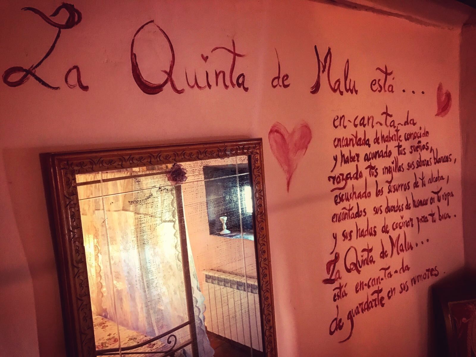 Un espejo con un marco decorativo refleja una habitación con ventana, un radiador y una inscripción en la pared.
