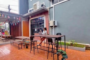 An outdoor seating area with tables and chairs, a person using a laptop, and a building with a grey facade. - You empire Hostel and Bar (Pakse)
