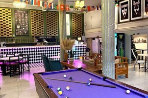 A billiards table in a room with a checkered wall, a bar area, and a seating area with chairs and tables.