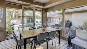 En täckt uteplats med ett träbord och stolar, en takfläkt och utsikt över en swimmingpool och ett hus.