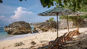 A beach with large rocks, a row of wooden beach chairs, and a striped umbrella. - The Korowai (Pecatu)