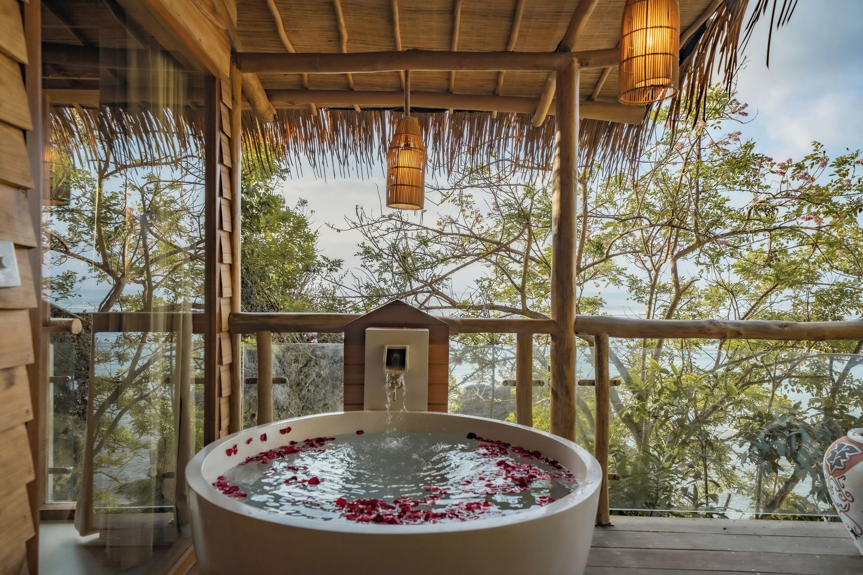 A wooden deck with a hot tub filled with water and rose petals, overlooking a natural landscape.