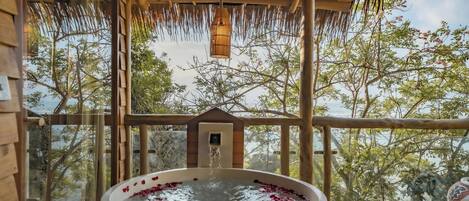 A wooden deck with a hot tub filled with water and rose petals, overlooking a natural landscape.