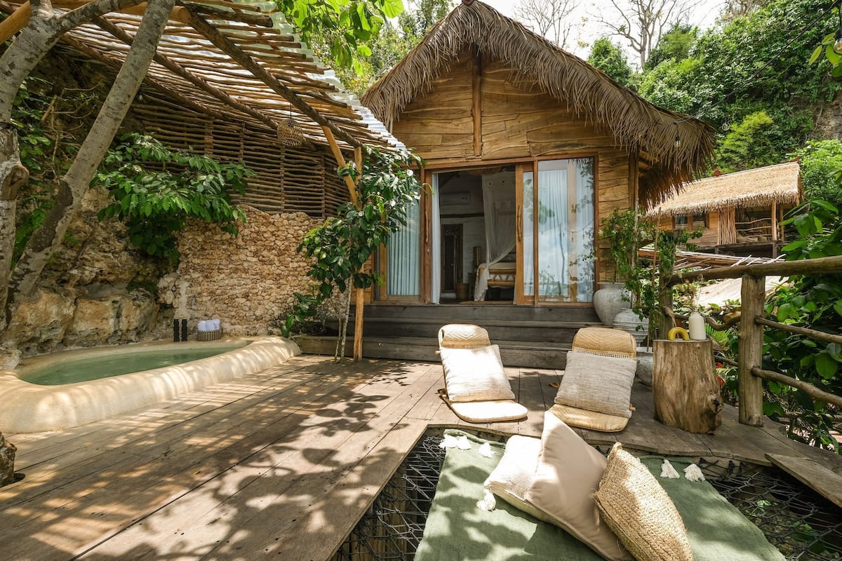 Una cabaña rústica de madera con techo de paja, pared de piedra y terraza de madera con sillones de descanso y una pequeña piscina.