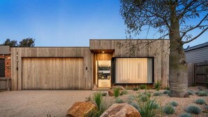 A modern house with a large garage, a well-maintained garden with rocks and plants, and a clear sky. - LUXE poolside living, in the heart of Flinders. (Flinders)