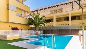 A swimming pool with a lifeguard chair, a palm tree, and a building with balconies. - Carnaval by Interhome (Isla Cristina)