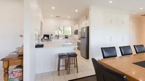 A modern kitchen with white cabinetry, a central island with stools, a dining area with a wooden table and black chairs, and a large refrigerator. - 131 Your Vineyard Home in the heart of the Vale (McLaren Flat)