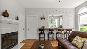 Sala de estar com lareira, mesa de centro de madeira com plantas em vasos, sofá de couro com almofada, e área de cozinha com banquetas e luzes de cordão.