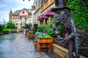 A cobblestone street with a statue of a man in a top hat and coat, a wooden cart with flowers, and buildings in the background. - Midea Egret Lake Car Theme Hotel (Foshan)