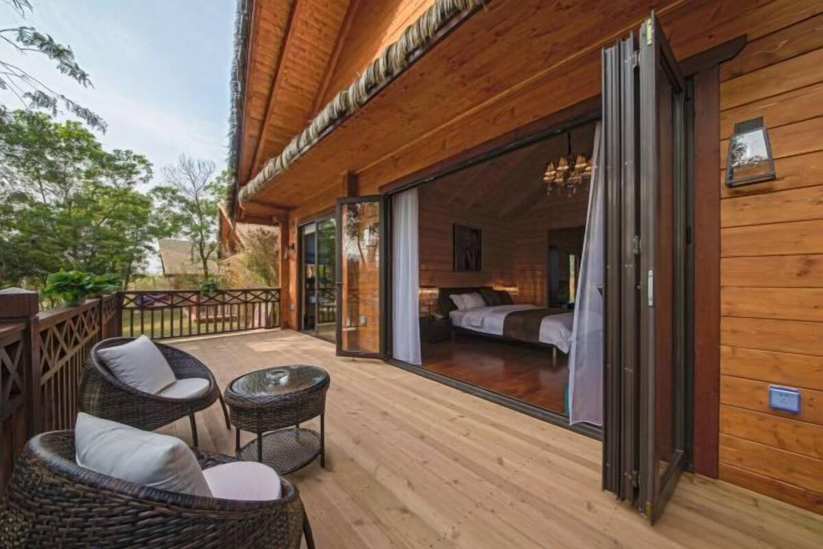 Una terraza de madera con muebles de mimbre y un ventanal que da a un dormitorio con cama y cortinas.