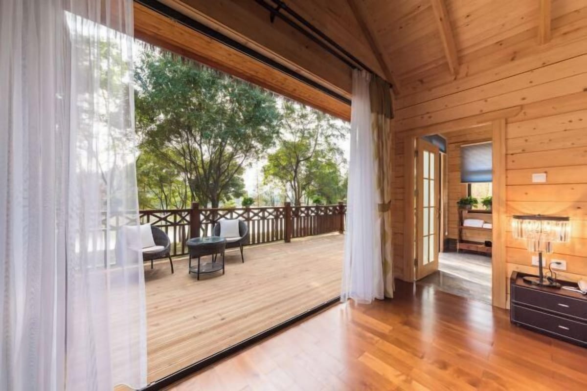 A wooden interior with a large window, a wooden deck outside, and a view of trees.