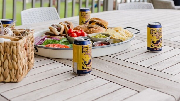 Een terras met eettafel, gedekt voor een maaltijd, inclusief een bord eten, een mand en twee blikjes bier.