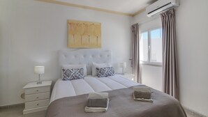 Ein ordentlich eingerichtetes Schlafzimmer mit Bett, Nachttischen, einer Wand-Klimaanlage und einem Fenster mit Vorhängen.