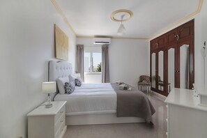 A bedroom with a bed, bedside table, dresser, wardrobe, and an air conditioning unit.
