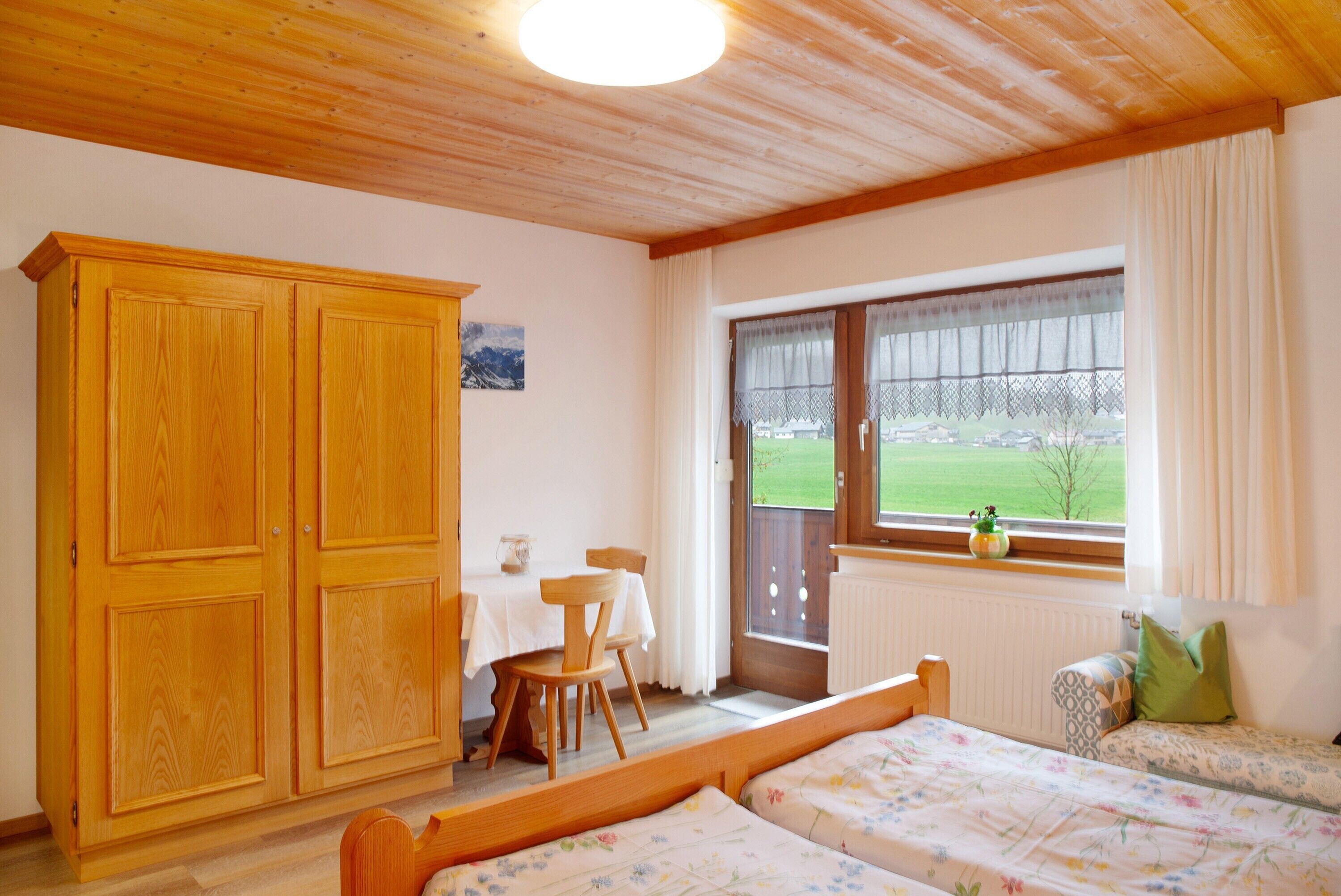A room with a wooden wardrobe, a bed with floral bedding, a table with chairs, and a window with a view of a green landscape.