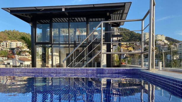 A modern pool area with a glass-enclosed structure, clear blue tiled pool, and a view of surrounding buildings. - A/C WIFI Zona Romantica Private Balcony Roof Top/Pool 5 Blocks from the beach (Puerto Vallarta)