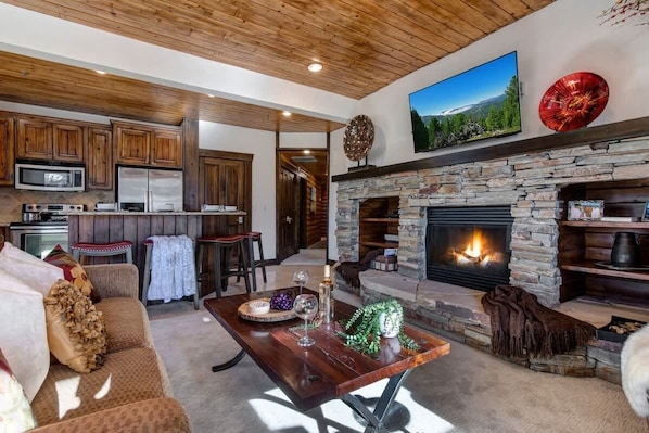 A spacious living area with a stone fireplace, wooden ceiling, and a kitchen in the background.