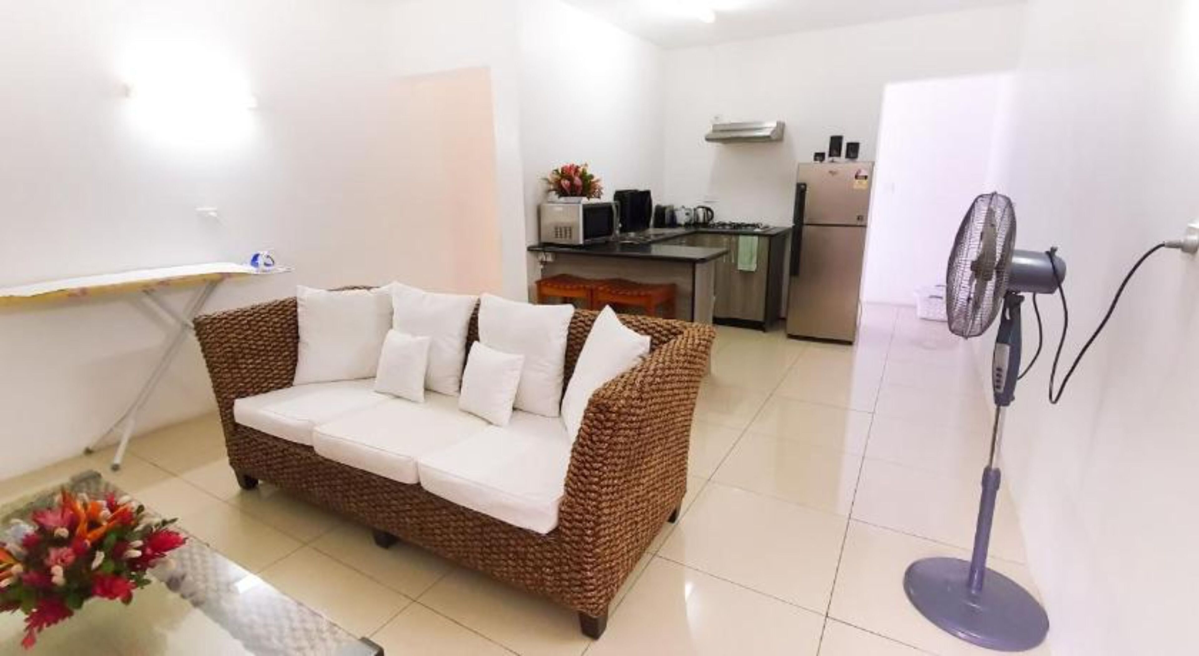 A living room with a brown wicker sofa, a standing fan, a kitchen area with a refrigerator, and a small table with flowers.