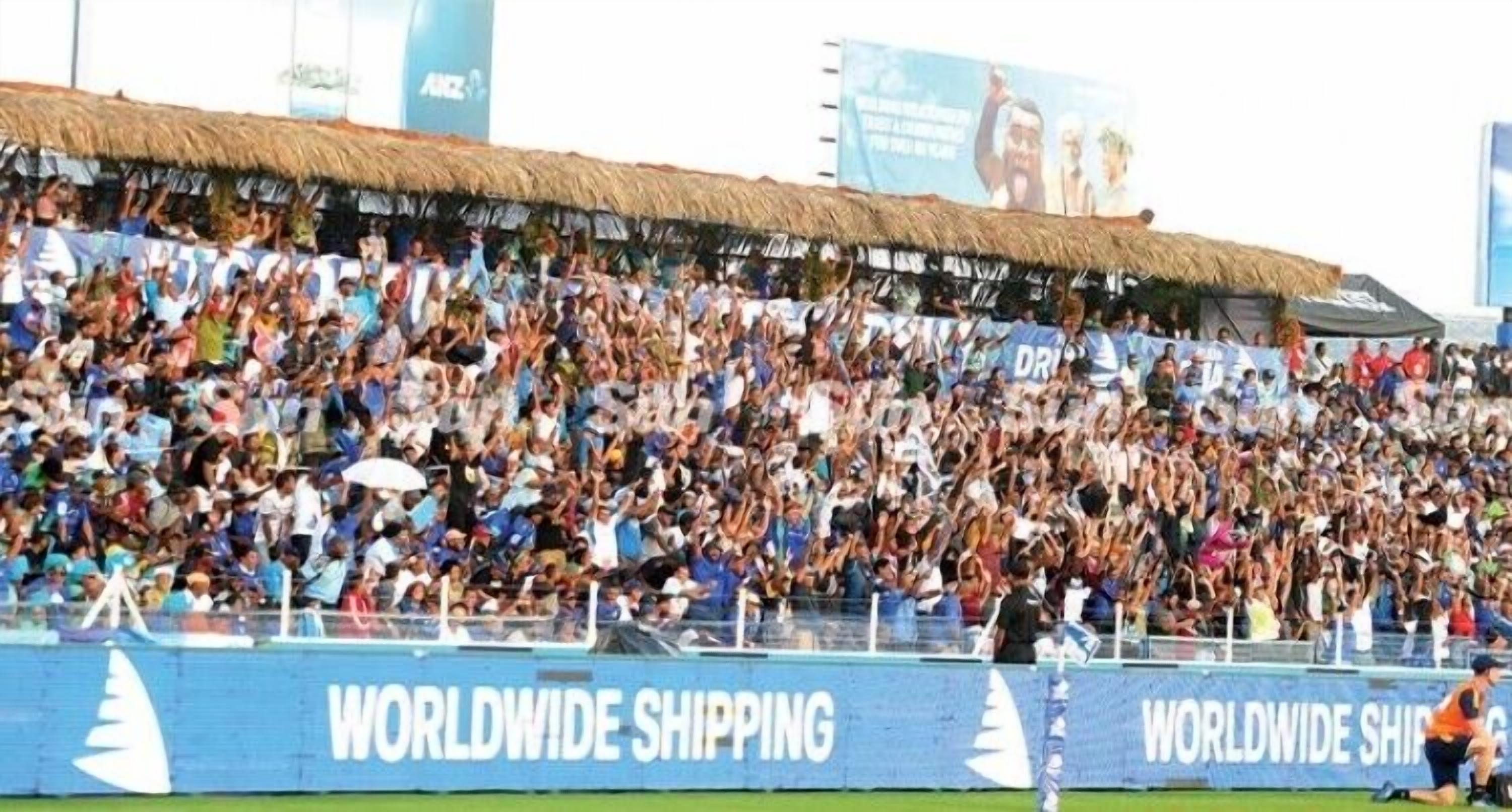 A stadium filled with spectators, with a thatched roof and advertisements for Worldwide Shipping.