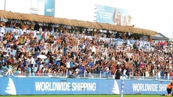 A stadium filled with spectators, with a thatched roof and advertisements for Worldwide Shipping.