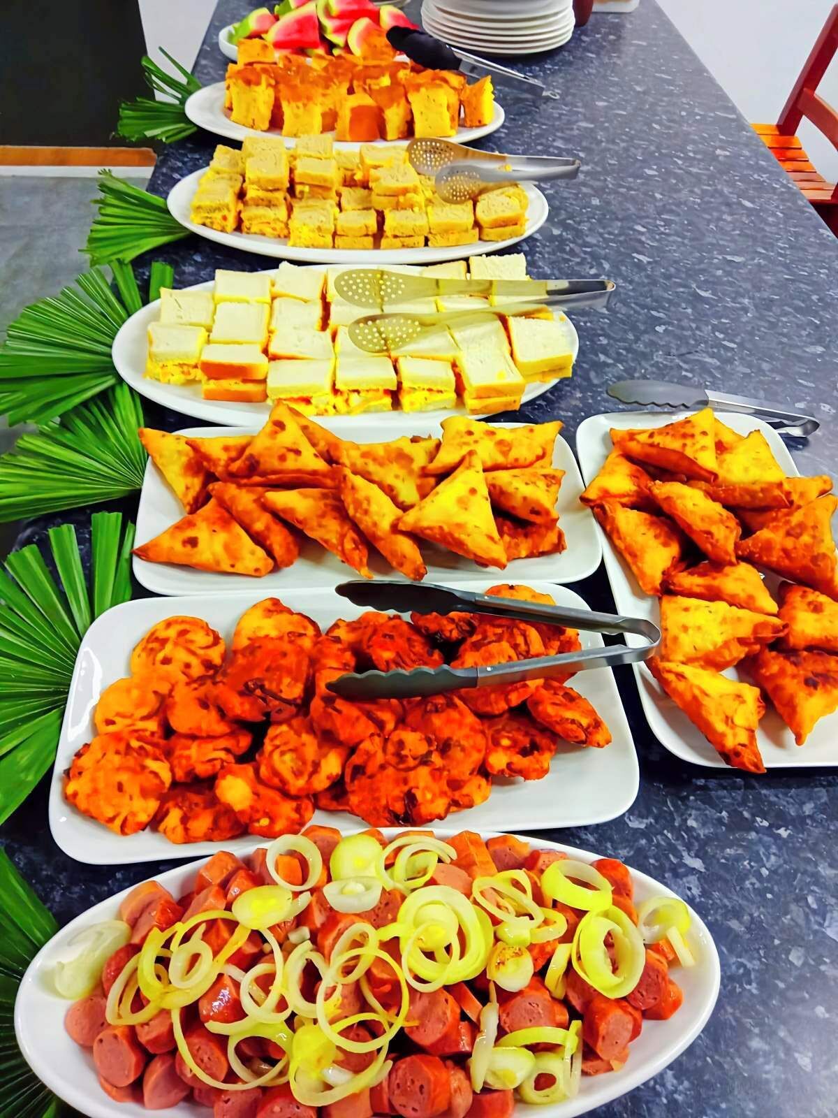 A buffet table with various dishes including omelettes, sandwiches, samosas, and a plate of sausages with onions.