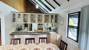 A dining area with a round table covered in a patterned tablecloth, surrounded by wooden chairs. A woven pendant light hangs from the ceiling, and the room features exposed wooden beams and a window with a view of greenery outside. - Island Accommodation Suva Premier Hospitality (Suva)