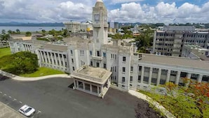 A large building with a prominent clock tower, surrounded by other structures and a body of water in the background. - Island Accommodation Suva Premier Hospitality (Suva)