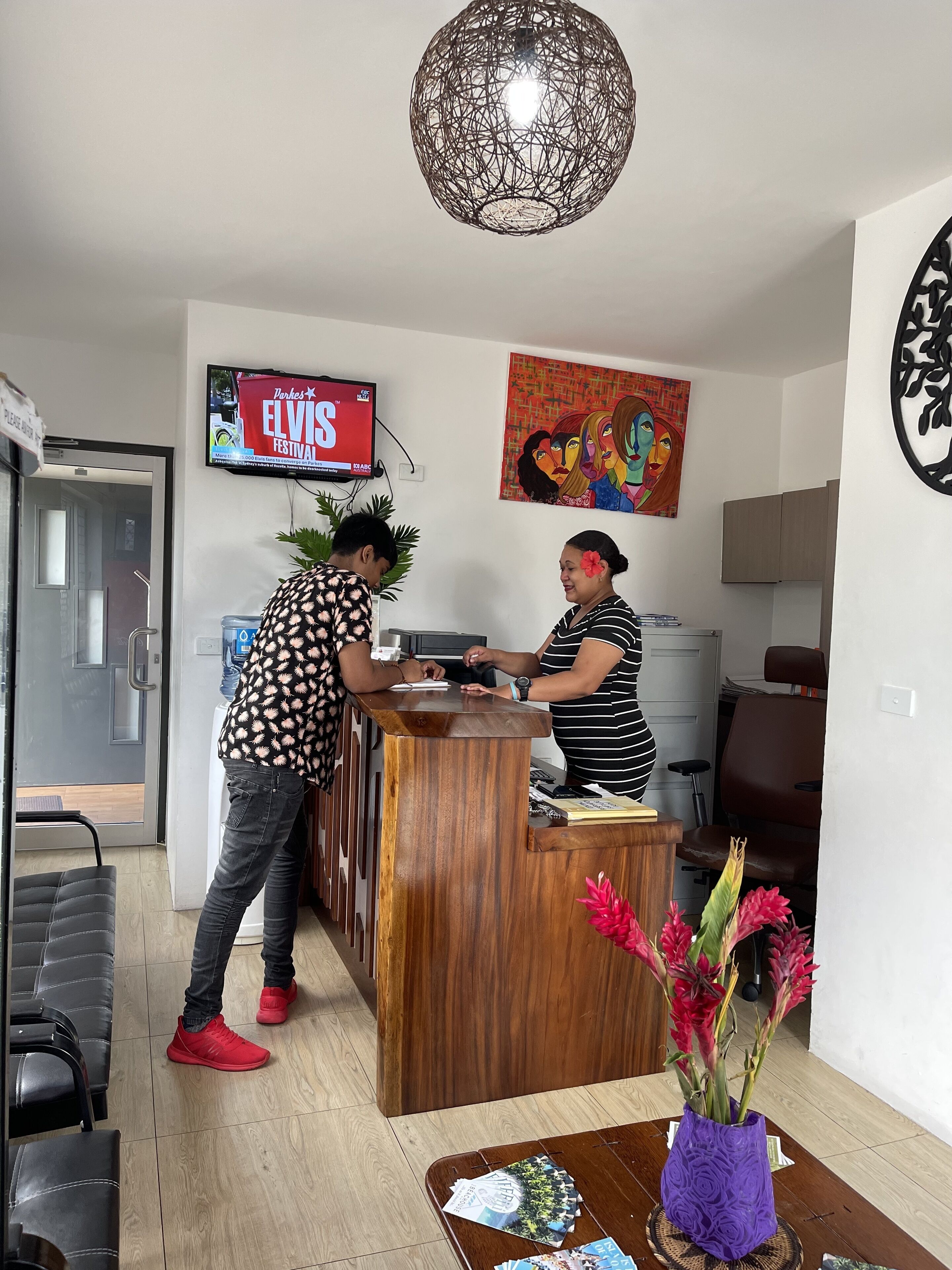 A reception desk with two staff members, a TV displaying 'Elvis Festival', and a colorful abstract painting on the wall.