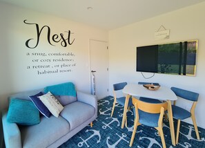 A cozy room with a grey sofa, blue cushions, and a small table with chairs. A TV is mounted on the wall, and there's a sign above it. - Peace and Tranquility ... farmland, river and west coast beach - perfection! (Mimi)