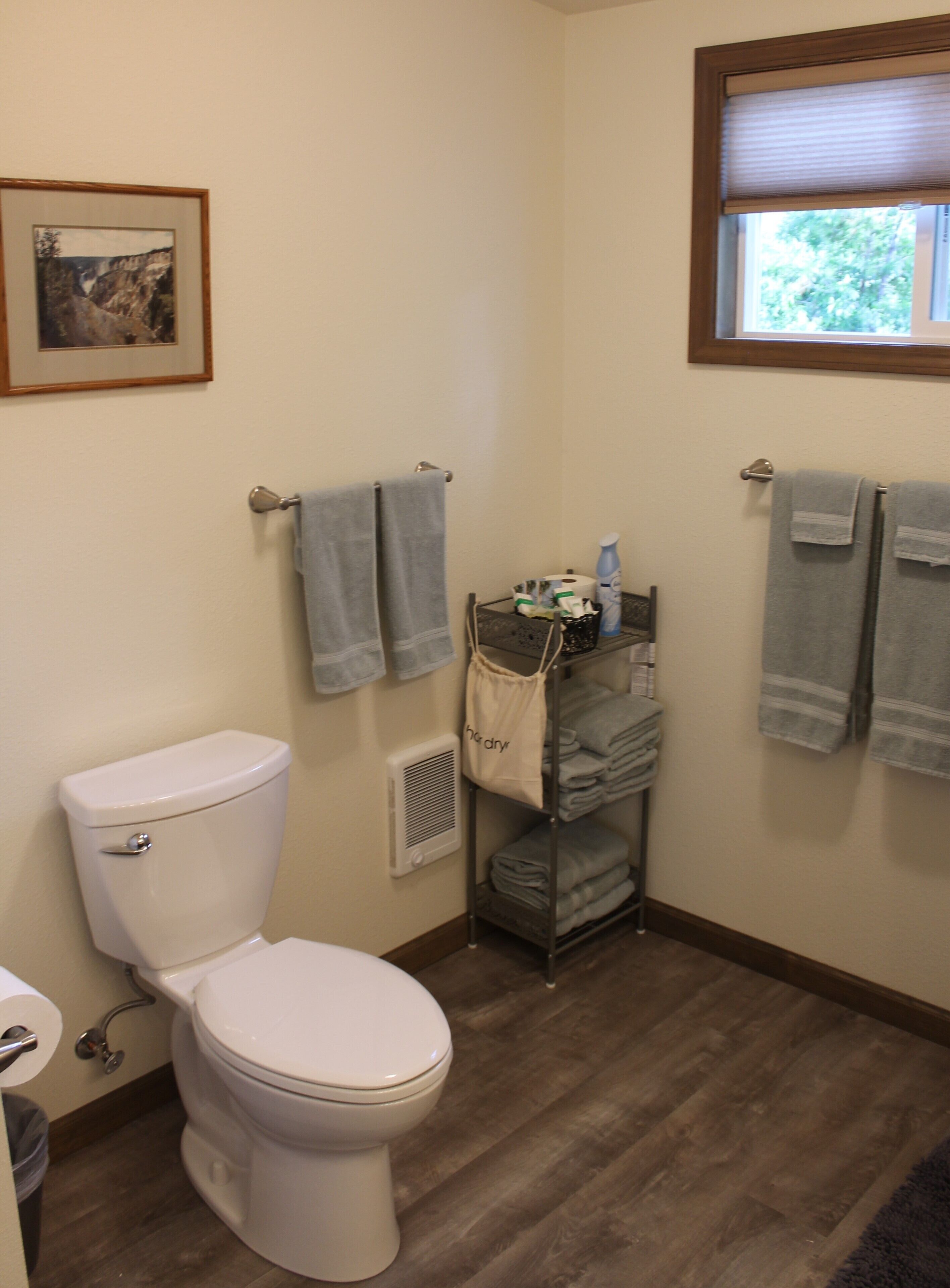 A bathroom with a toilet, a towel rack with towels, a shelf with toiletries, and a framed picture on the wall.