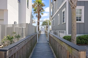 A wooden boardwalk leading to a gated entrance with a palm tree and a building with a gray facade.