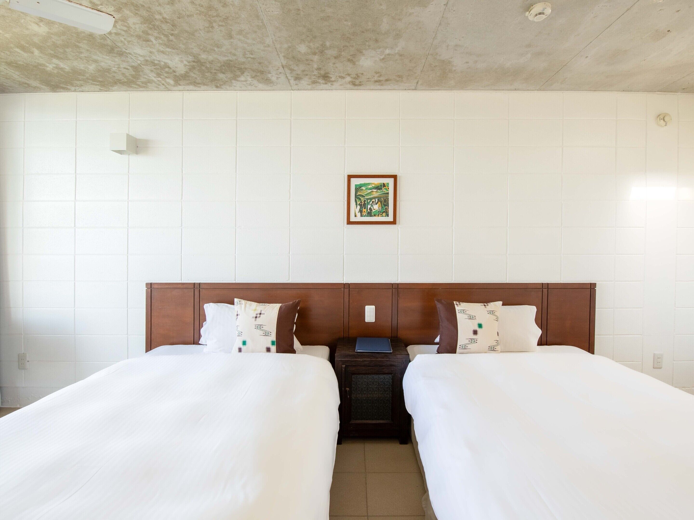 Een hotelkamer met twee enkele bedden, een houten hoofdbord, witte beddengoed, een klein nachtkastje met een lamp, en een lijst met een afbeelding aan de muur.