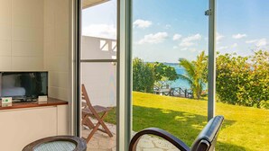 A room with a TV on a wooden table, a stone bench, and a glass door leading to a patio with a view of the sea and greenery. - 413 Hamahiga Hotel&cafe (Uruma)