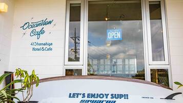 Una tabla de surf con el texto "LET'S ENJOY SUP!!" y "SAKURA", colocada frente a un edificio con un letrero que dice "Oceanview Cafe 413 hama higa Hotel & Cafe" y un letrero "ABIERTO" sobre la entrada.