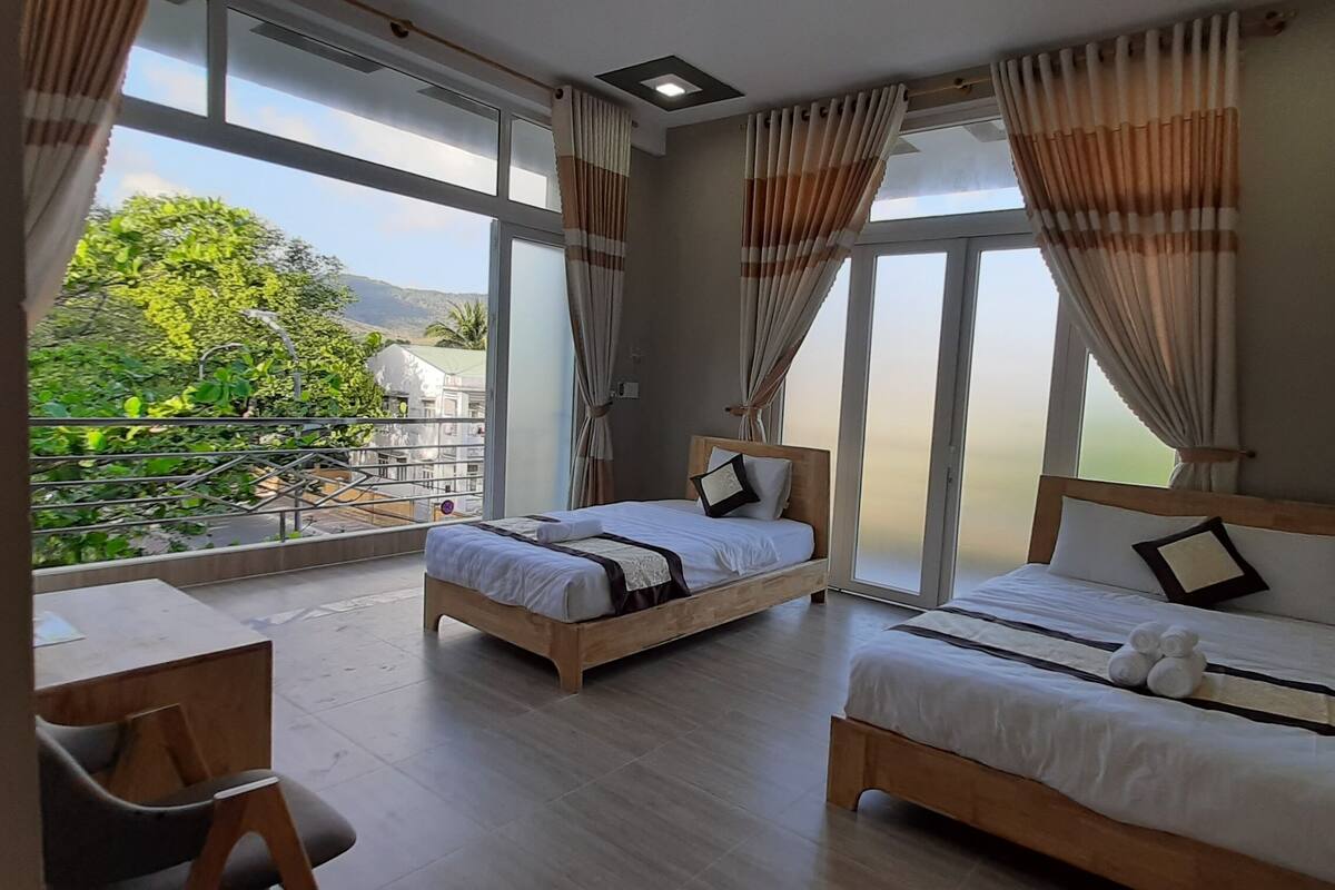 A hotel room with two beds, a balcony with a view of greenery and mountains, and a wooden desk.