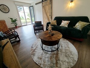 Una sala de estar con una mesa redonda, un sofá y una silla. Hay una planta en el suelo y una puerta que da al exterior.