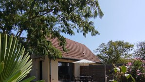 Een huis met een pannendak, een terras met tuinmeubilair en een tuin met planten en een rode bloem.