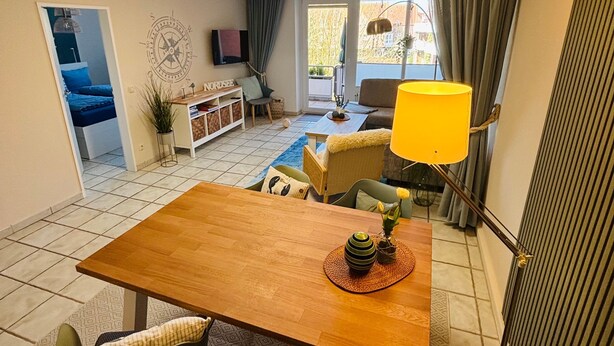 A dining area with a wooden table, chairs, and a yellow lamp. There is a small plant on the table and a view of a balcony with a chair and a TV in the background.