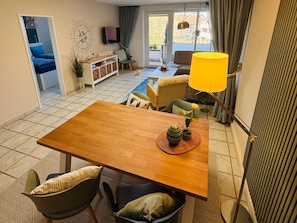 A dining area with a wooden table, chairs, and a yellow lamp. There is a small plant on the table and a view of a balcony with a chair and a TV in the background. - Atlanta 21 - 70006, Deutschland (Wangerland)