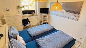 A bedroom with a bed, two hanging lamps, a TV, and a window with a view of a house.
