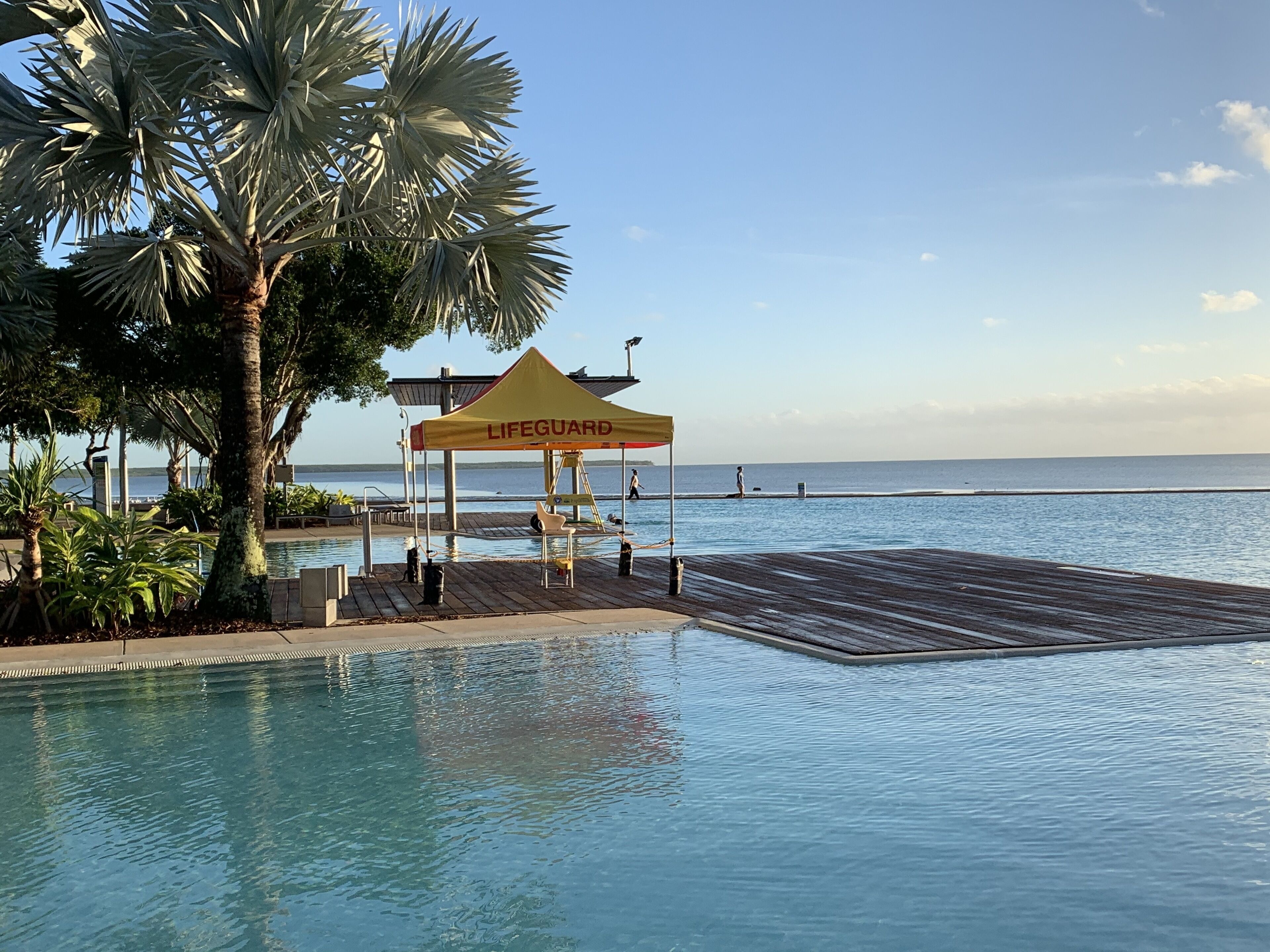 Una estación de salvavidas junto a una piscina con muelle de madera y palmeras.
