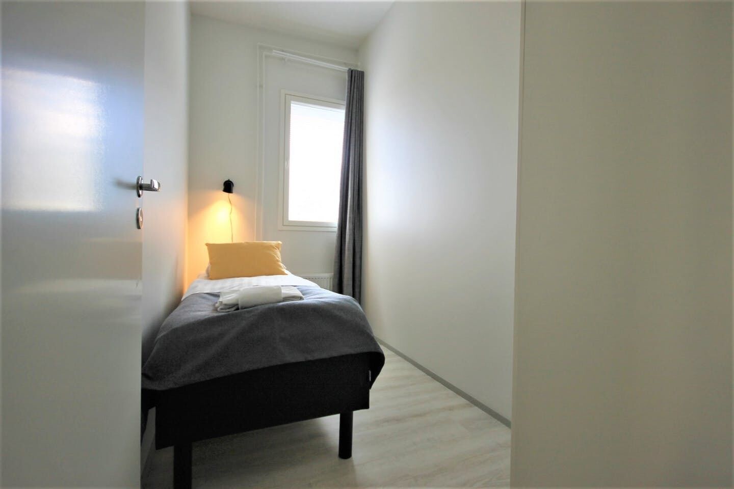 Una habitación estrecha y bien iluminada, con una cama, una ventana con cortinas y una puerta.