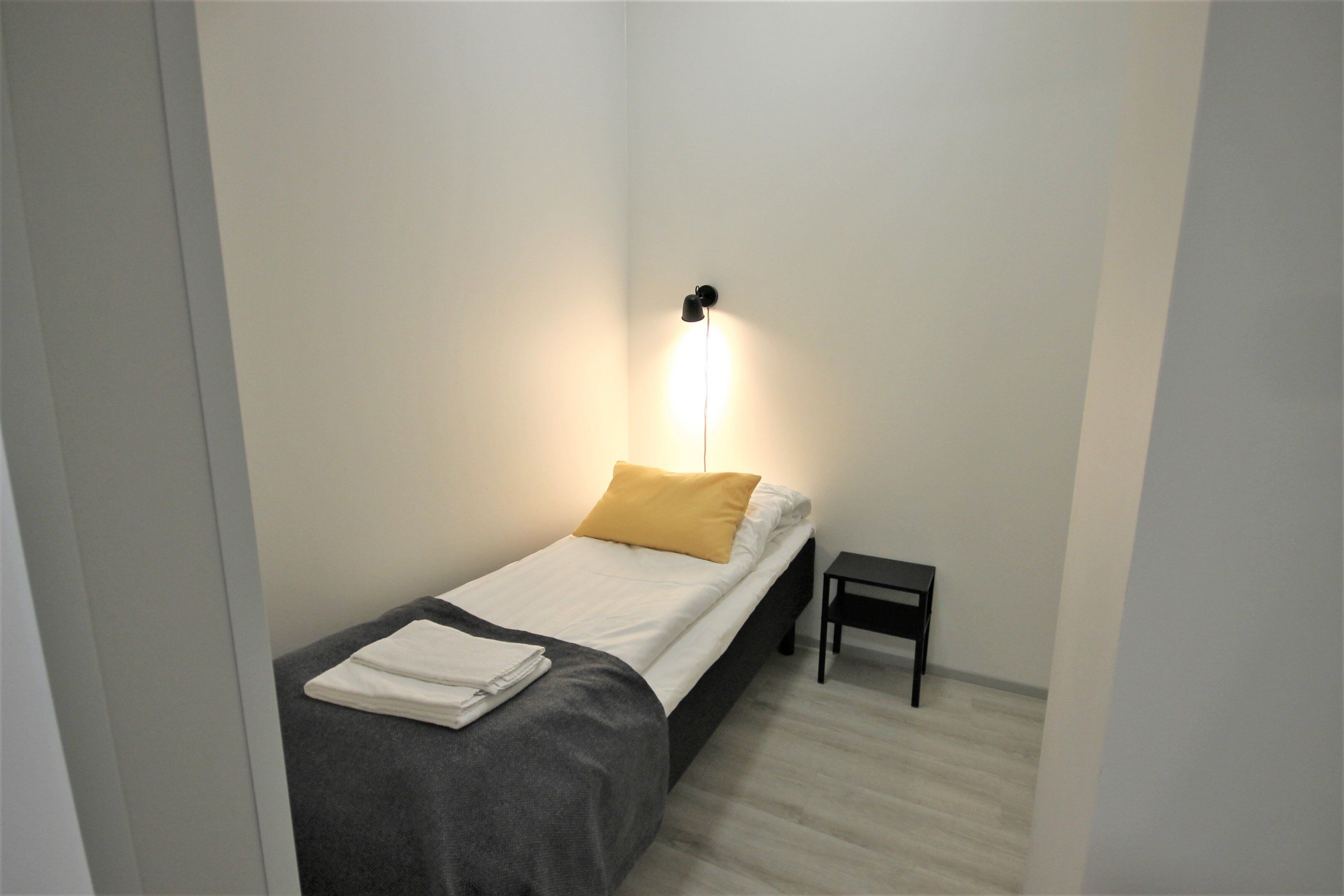A small, neatly made bed with a white and gray bedspread, a yellow pillow, and a folded white towel on the side. There is a small black nightstand beside the bed.