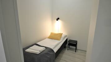 A small, neatly made bed with a white and gray bedspread, a yellow pillow, and a folded white towel on the side. There is a small black nightstand beside the bed.