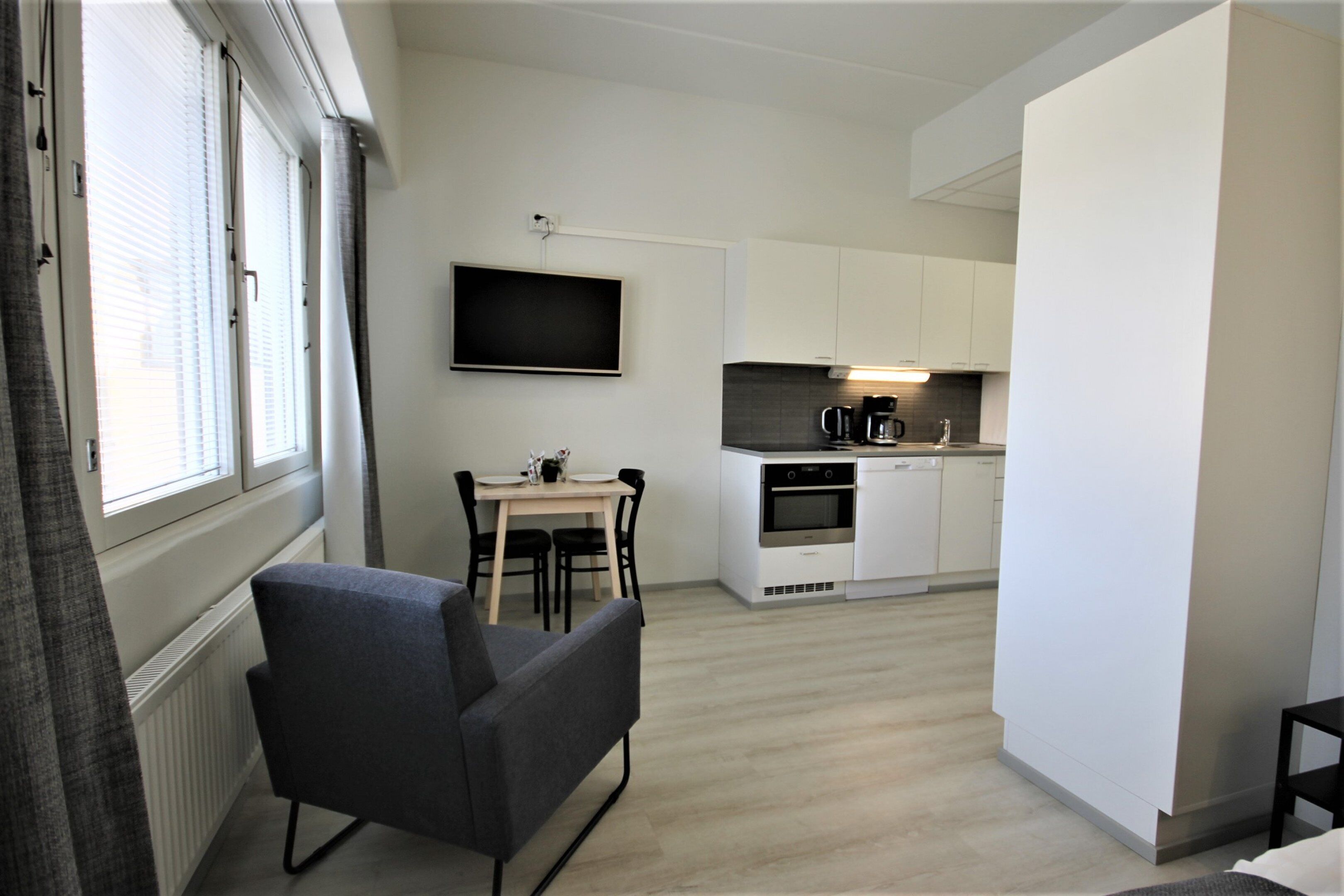 Een moderne keuken met witte kastjes, een ingebouwde oven en een koffiezetapparaat. Er is een zithoek met een tafel en stoelen, en een grijze bank.