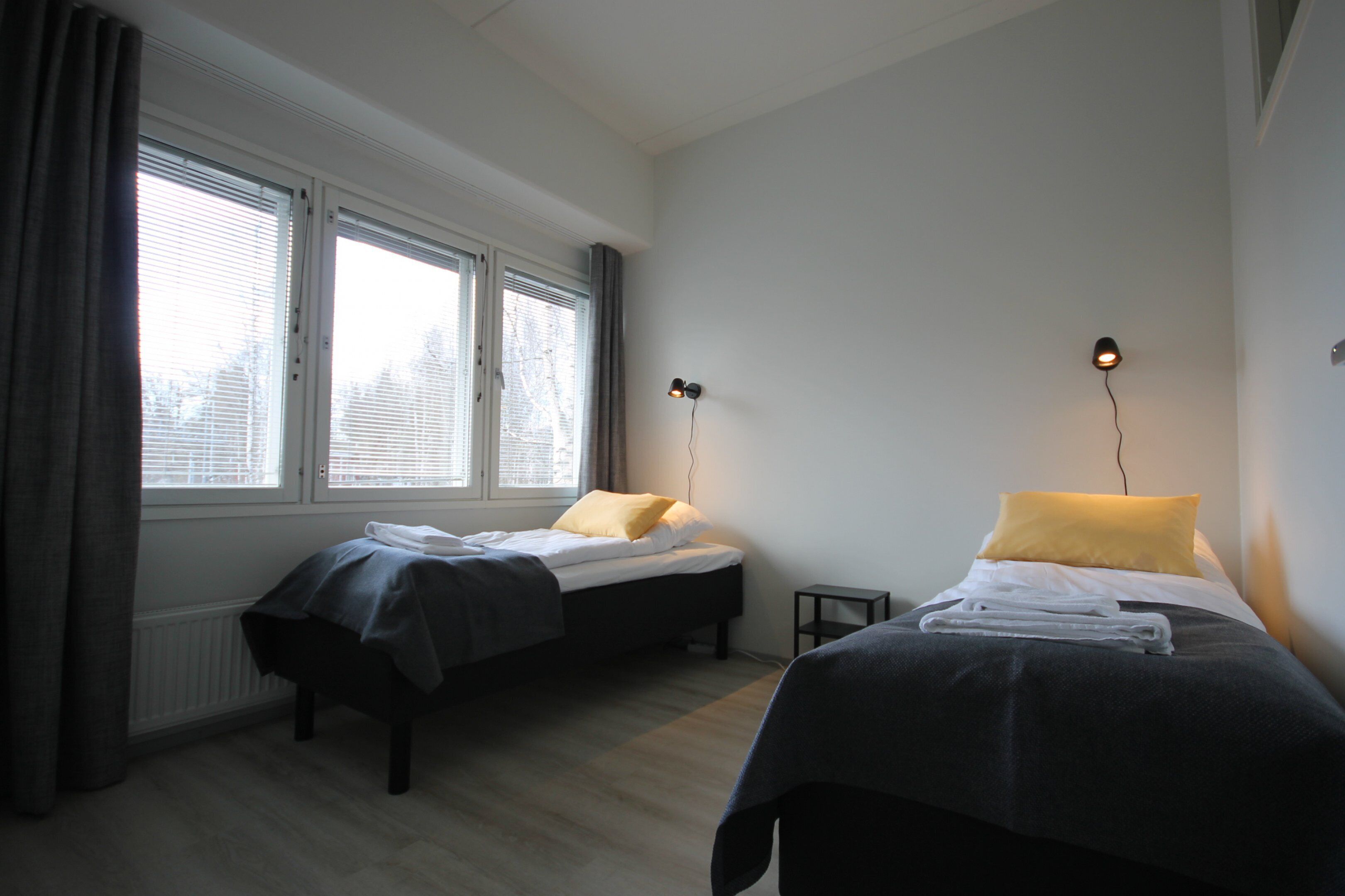 A room with two beds, a window with blinds, a small table, and a wall-mounted lamp.